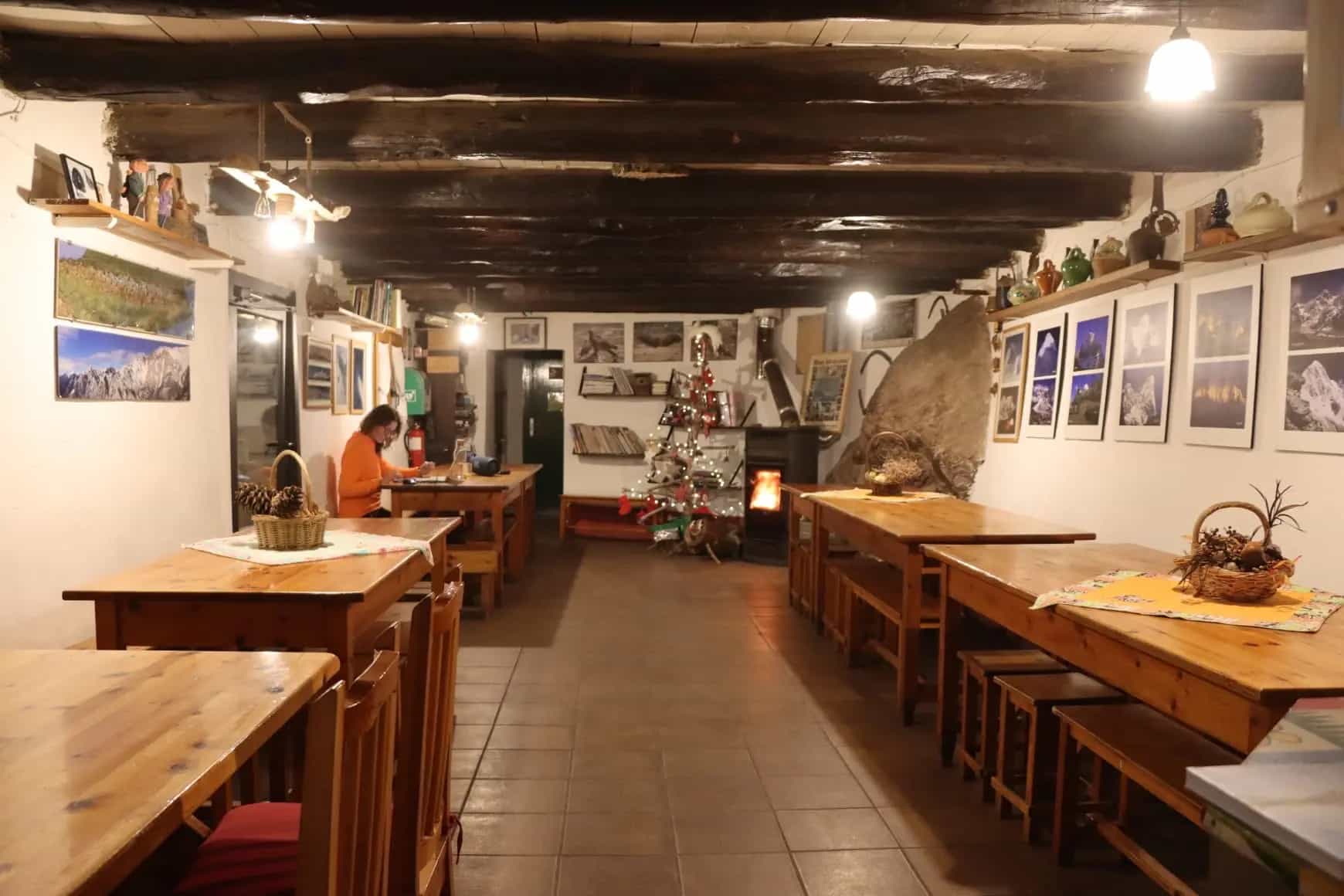 Rustic dining hall with wooden tables, exposed beams, a small Christmas tree, and a wood stove.