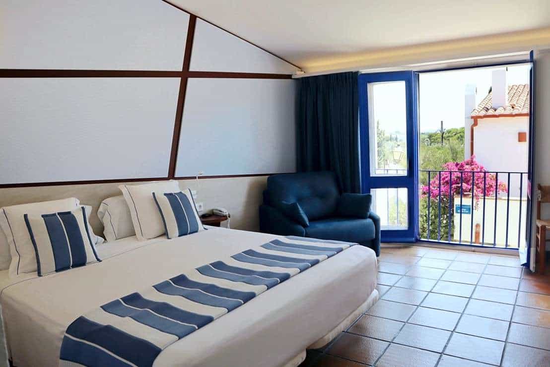 Hotel room with white and blue bedding, blue armchair, and balcony view of white building.