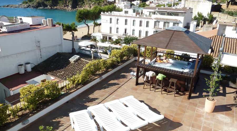 Rooftop hot tub with two people, overlooking beach and coastal town buildings.