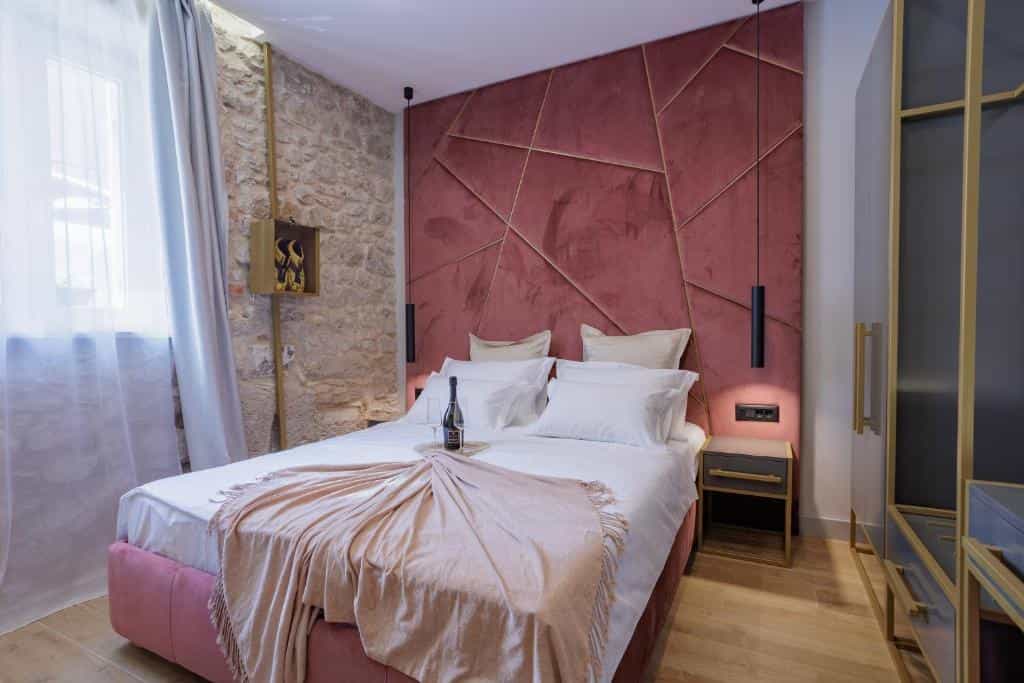Bedroom with pink velvet headboard, stone wall, white linens, and champagne bottle on bed.