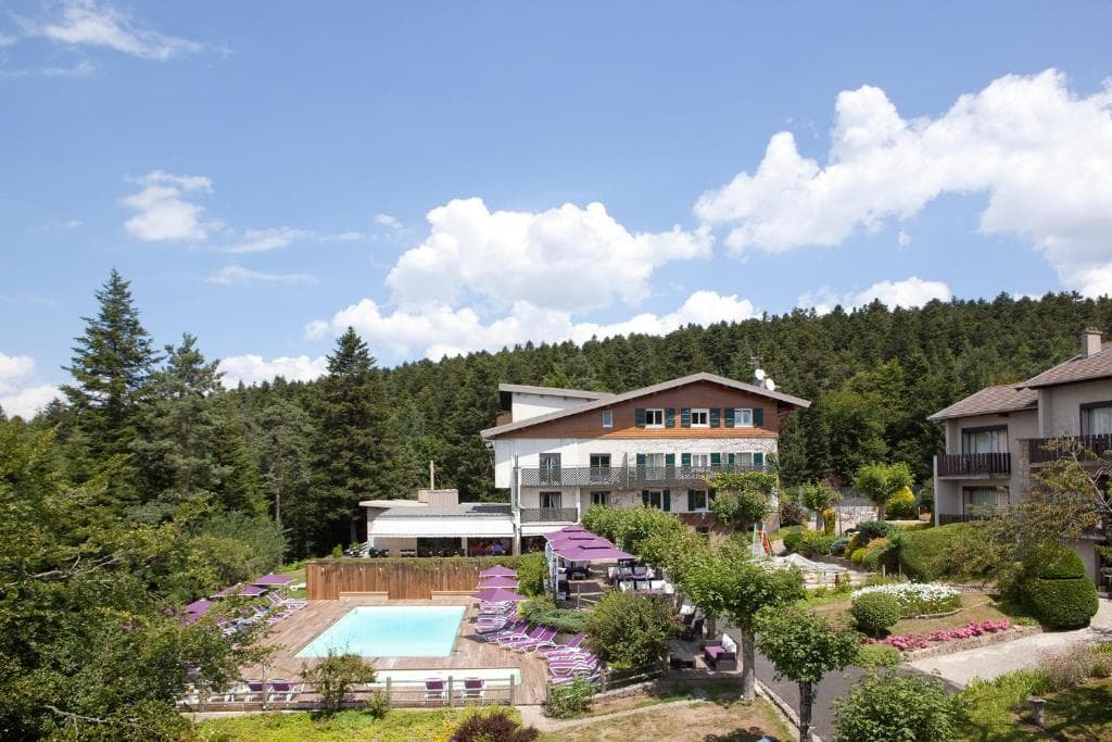 Hotel with outdoor pool and sun loungers nestled against a dense pine forest under a blue sky.