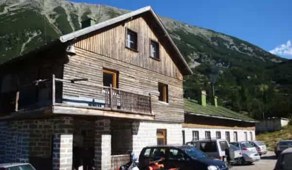 Wooden mountain hut with stone base and parking lot against a steep, forested mountain.