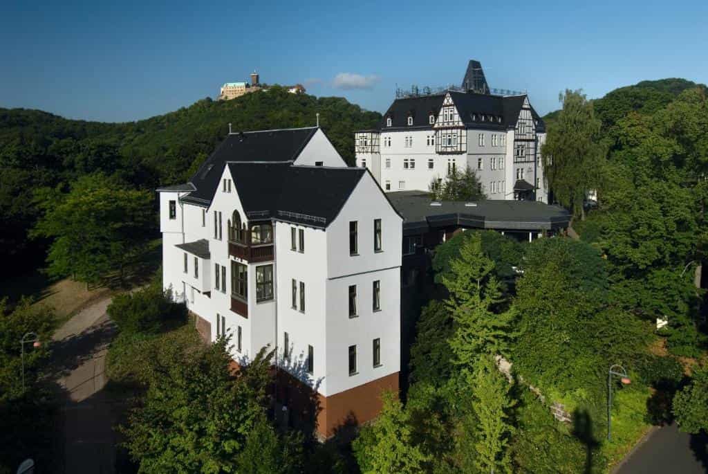 White buildings with black roofs nestled in green hillside trees, castle visible on distant hill.