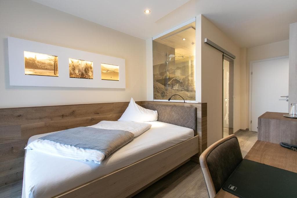 Hotel room with wooden bed frame, white linens, and mountain-themed artwork near glass shower enclosure.