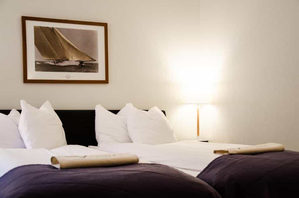 Hotel room with twin beds, white pillows, and framed sailboat artwork above the headboard.