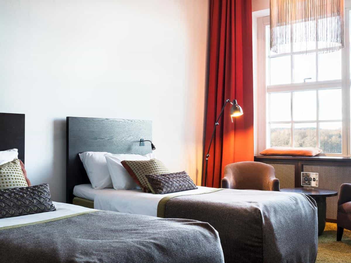 Twin beds in a hotel room with white walls, red curtains, and a view of trees outside the window.