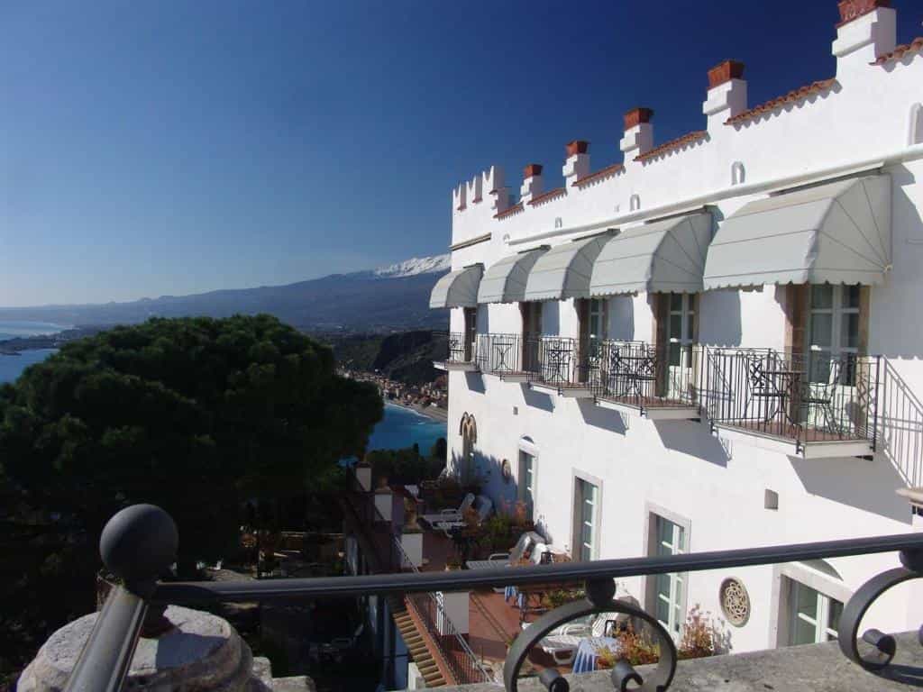 White hotel exterior with balconies overlooking a coastal town and snow-capped mountain.