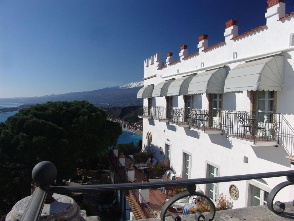 White hotel exterior with balconies overlooking a coastal town and snow-capped mountain.