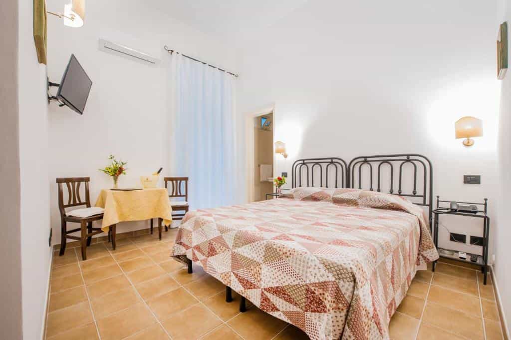 Bright hotel bedroom with metal-frame bed, patterned quilt, and small dining table.