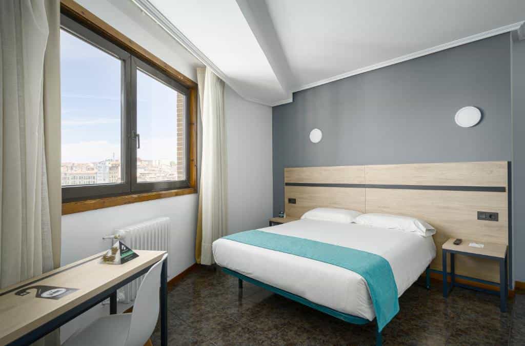 Hotel room with double bed, desk, and city view through large window.