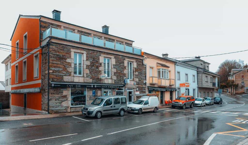 Cafe-Bar Mesón a Marronda building with stone facade and orange side on wet street with parked cars.