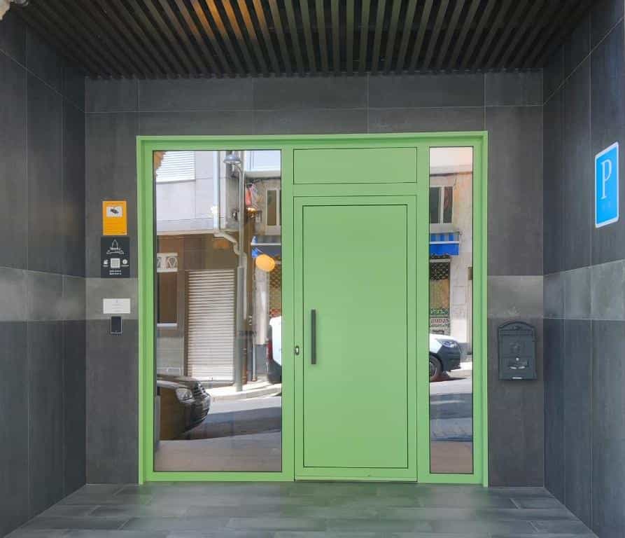 Entrance with bright green door and glass panels set in dark gray tiled wall, reflecting a street.