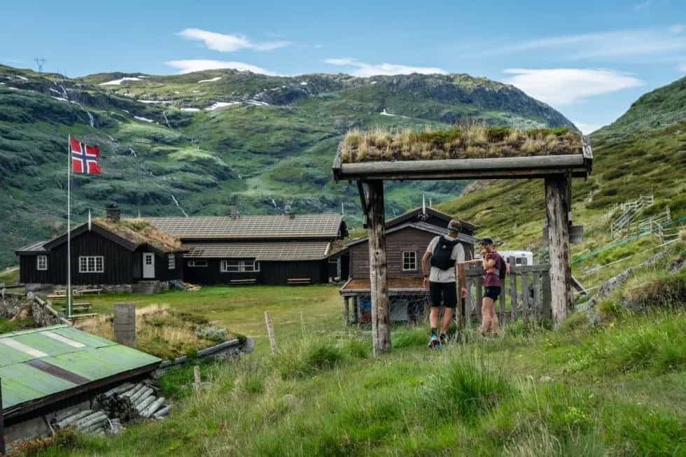 Hikers at Steinbergdalshytta mountain lodge with a Norwegian flag and green mountain landscape.