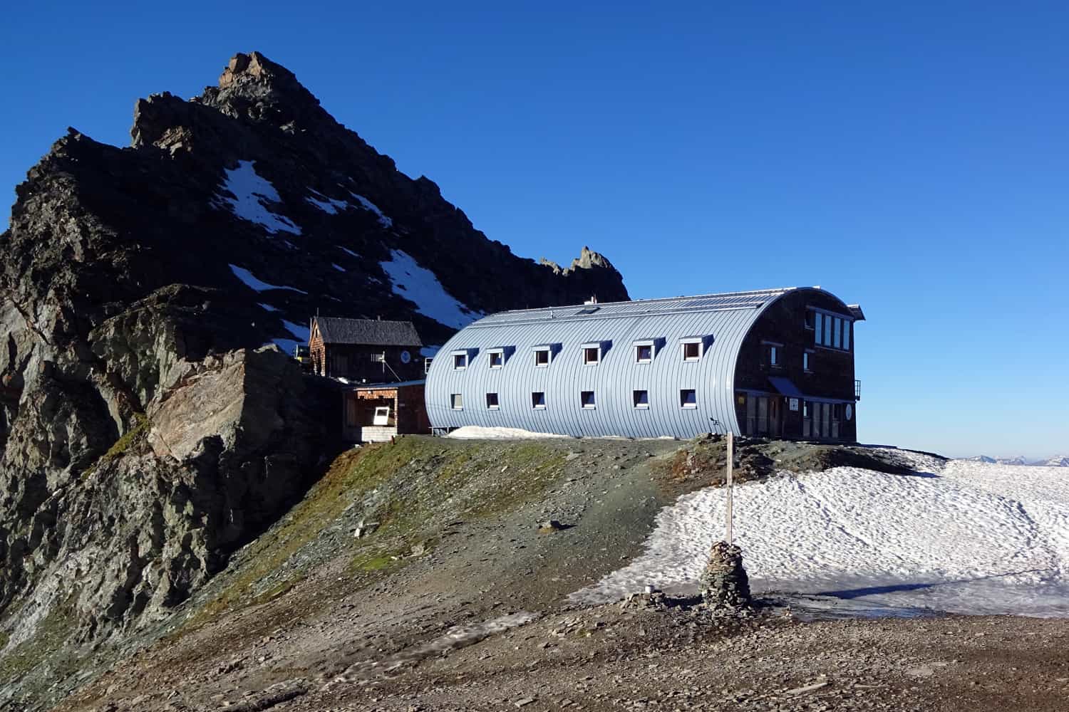 Stüdlhütte mountain hut with curved metal roof beside rocky peak and patches of snow under clear blue sky.