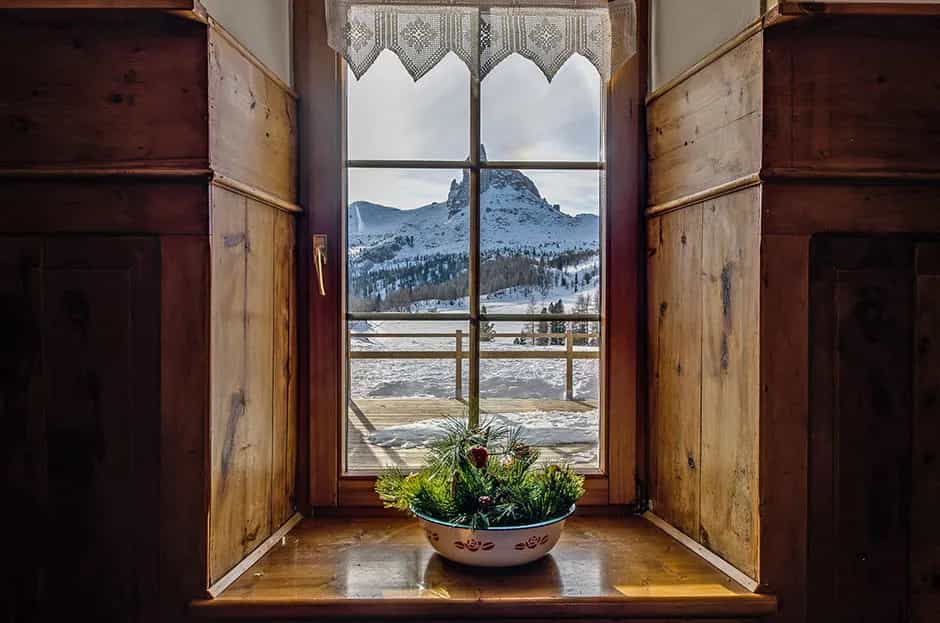Window view of Rifugio Croda da Lago with snow-covered mountains and pine centerpiece.
