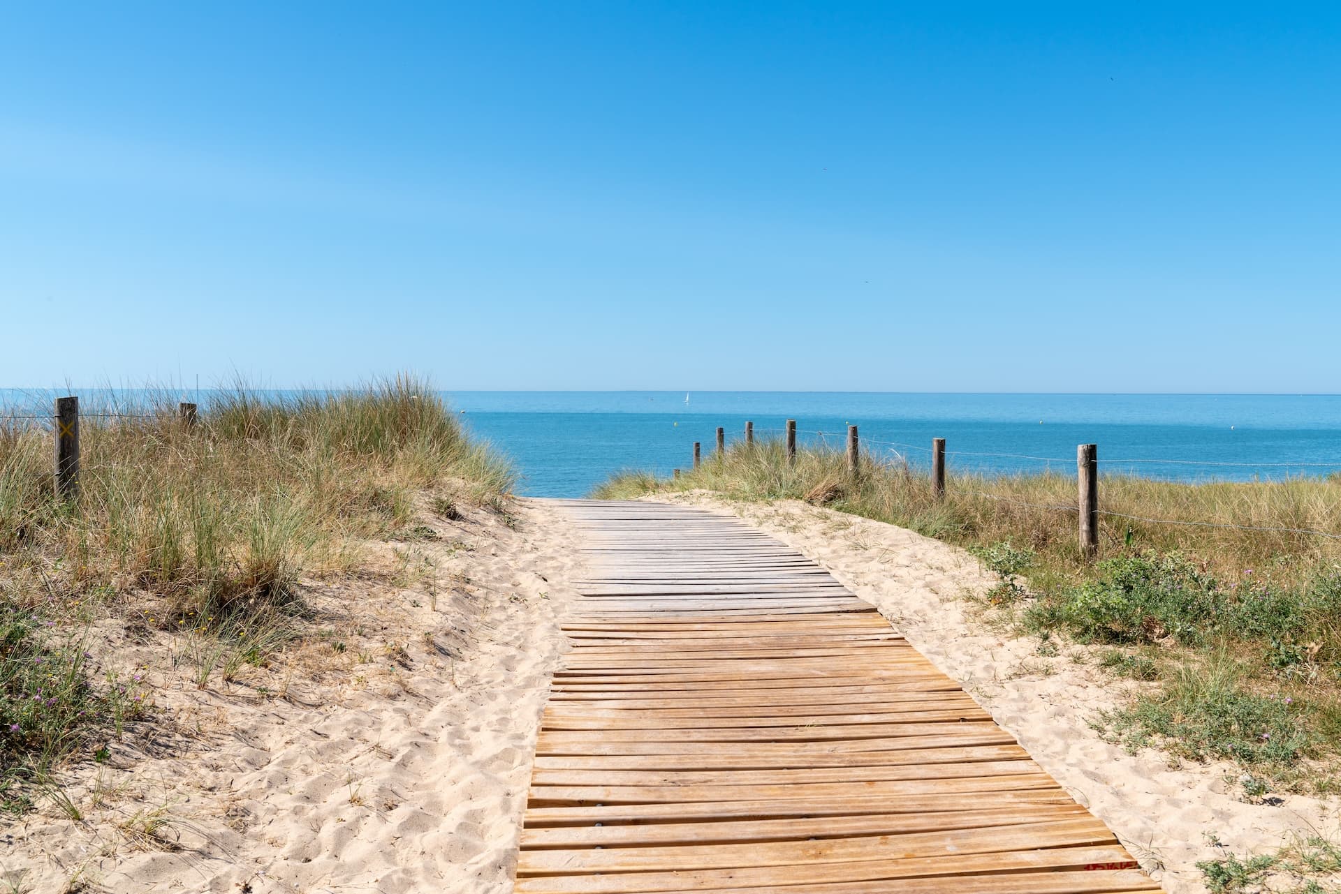 Wooden boardwalk through sand dunes leading to the bright blue sea under a clear sky.