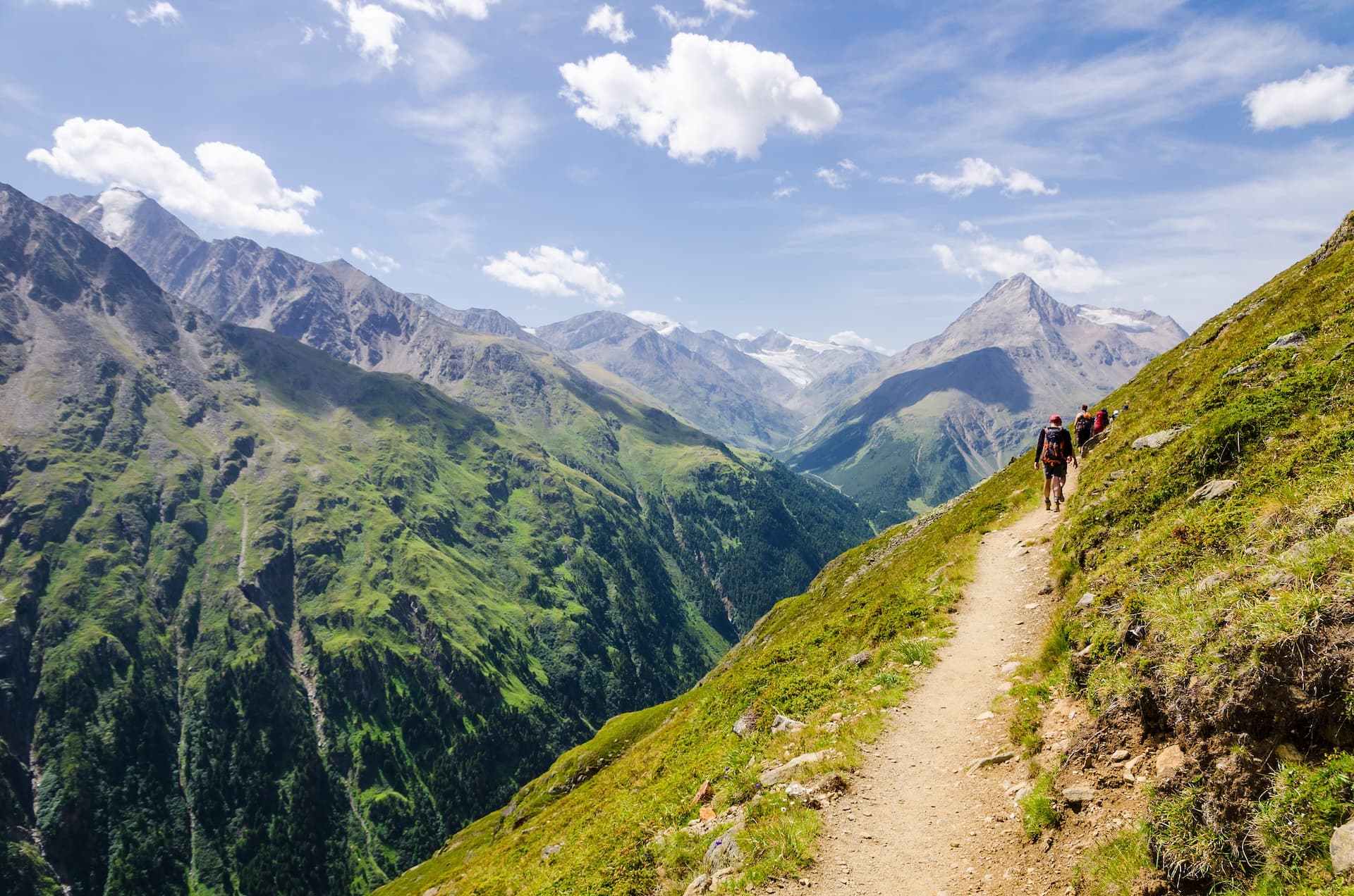 Hikers trekking on narrow dirt path along steep grassy slope with massive alpine mountains in background