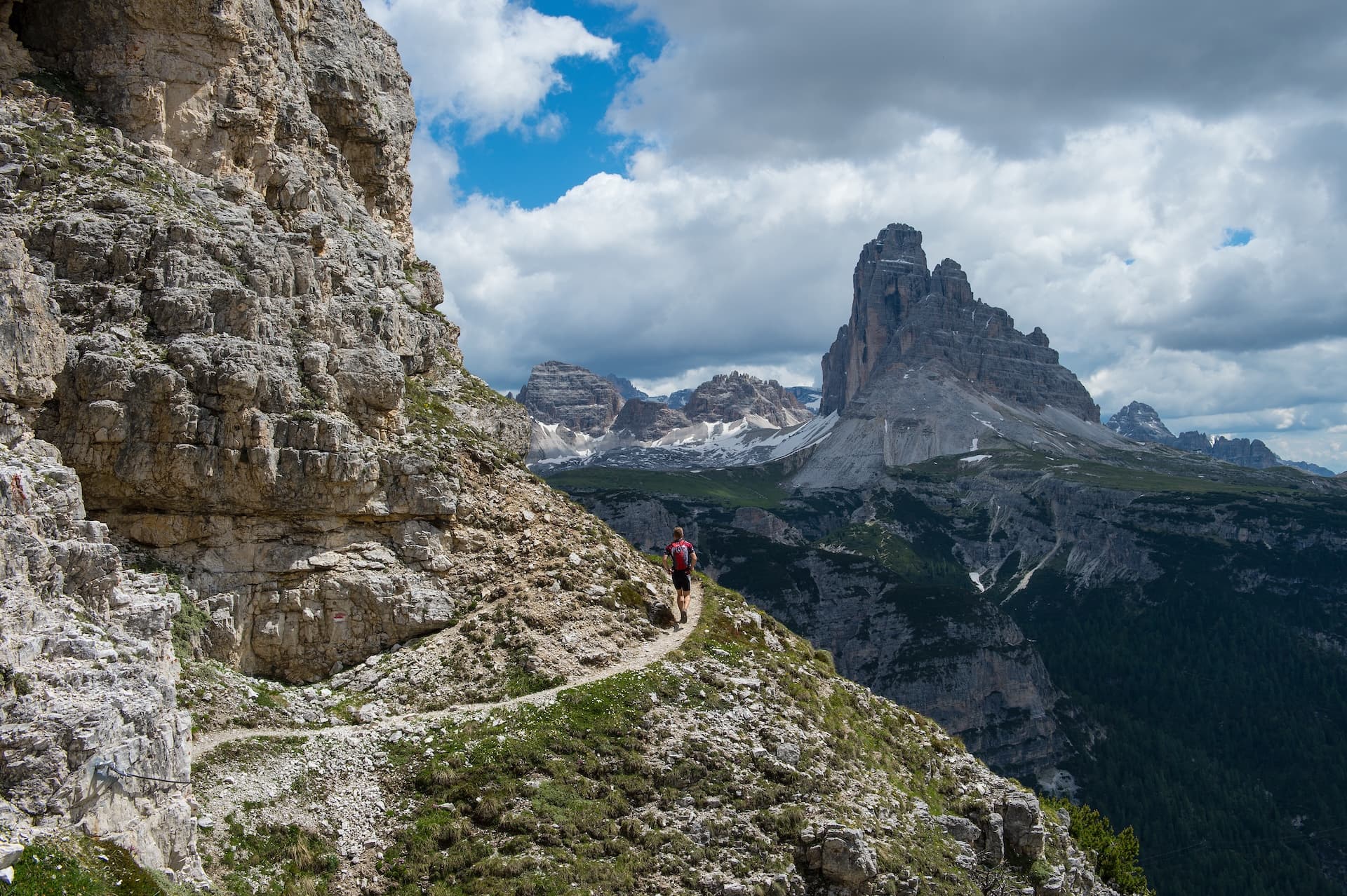 Hiker on narrow trail along rocky mountainside with dramatic peaks in background