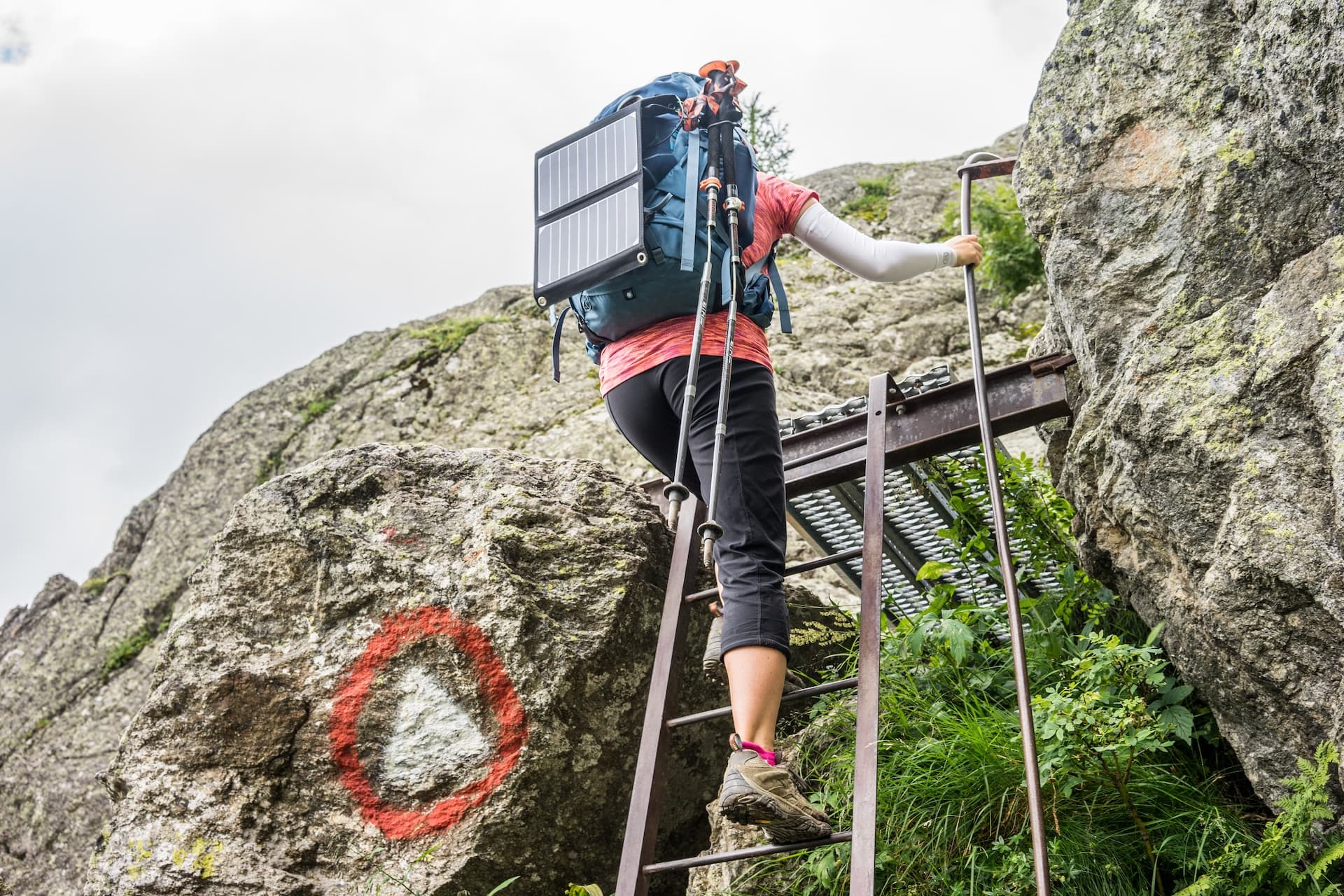 Hiker with solar panel backpack ascending metal ladder on rocky mountain trail with trail marker.