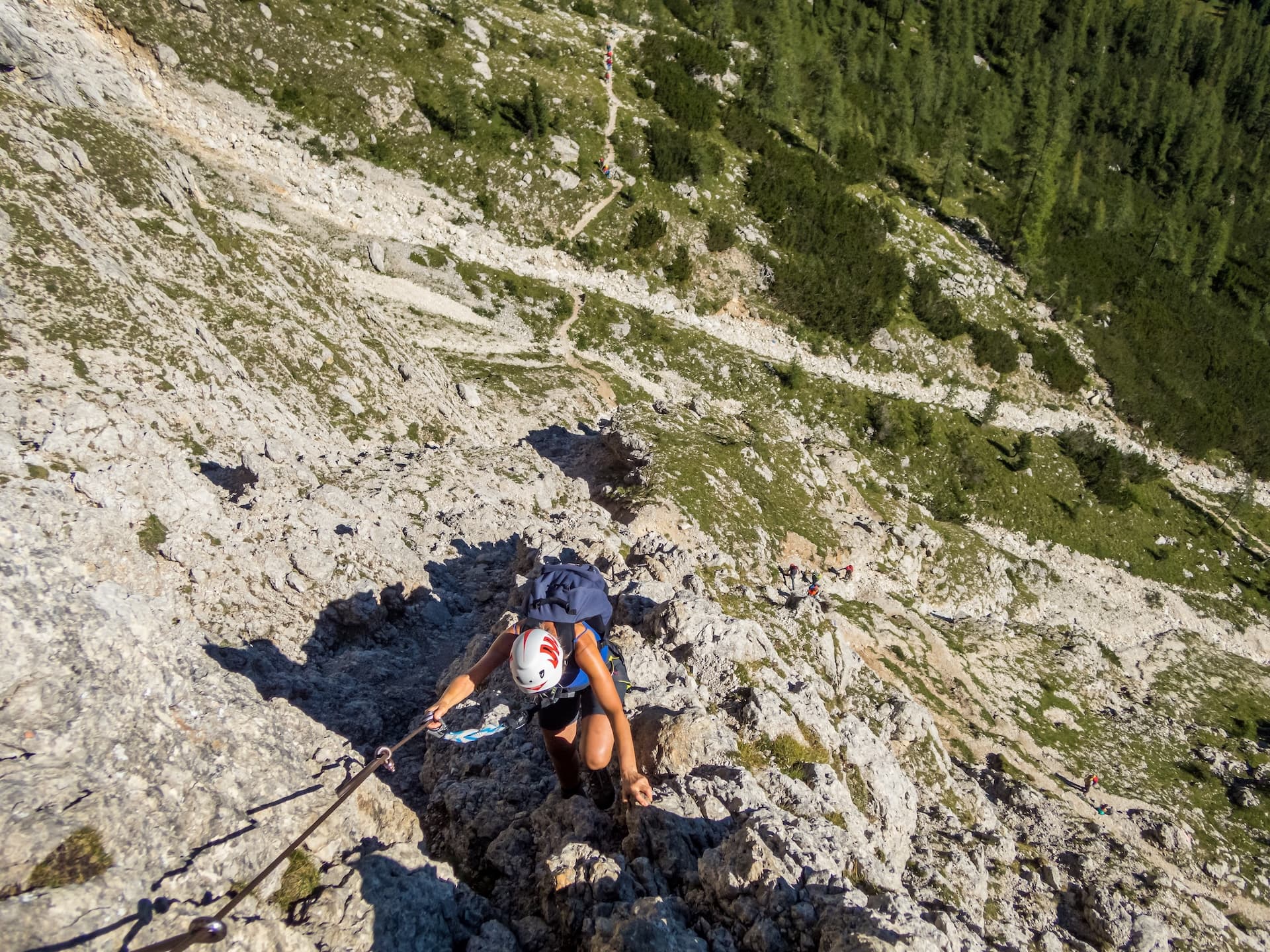 Woman climbing steep rocky mountain path using fixed cable, hikers visible below
