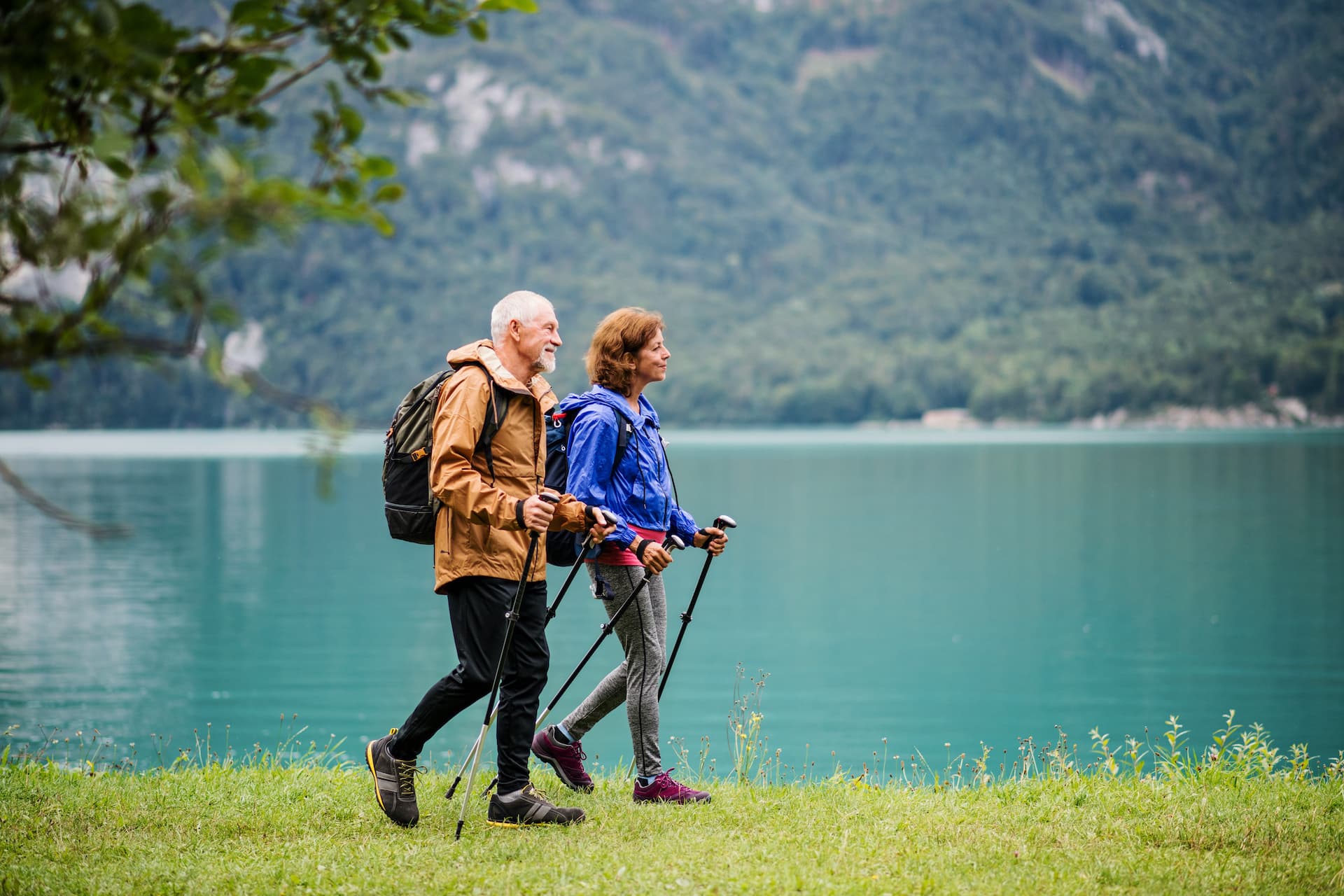 Senior couple hiking with poles on grassy shore by turquoise mountain lake