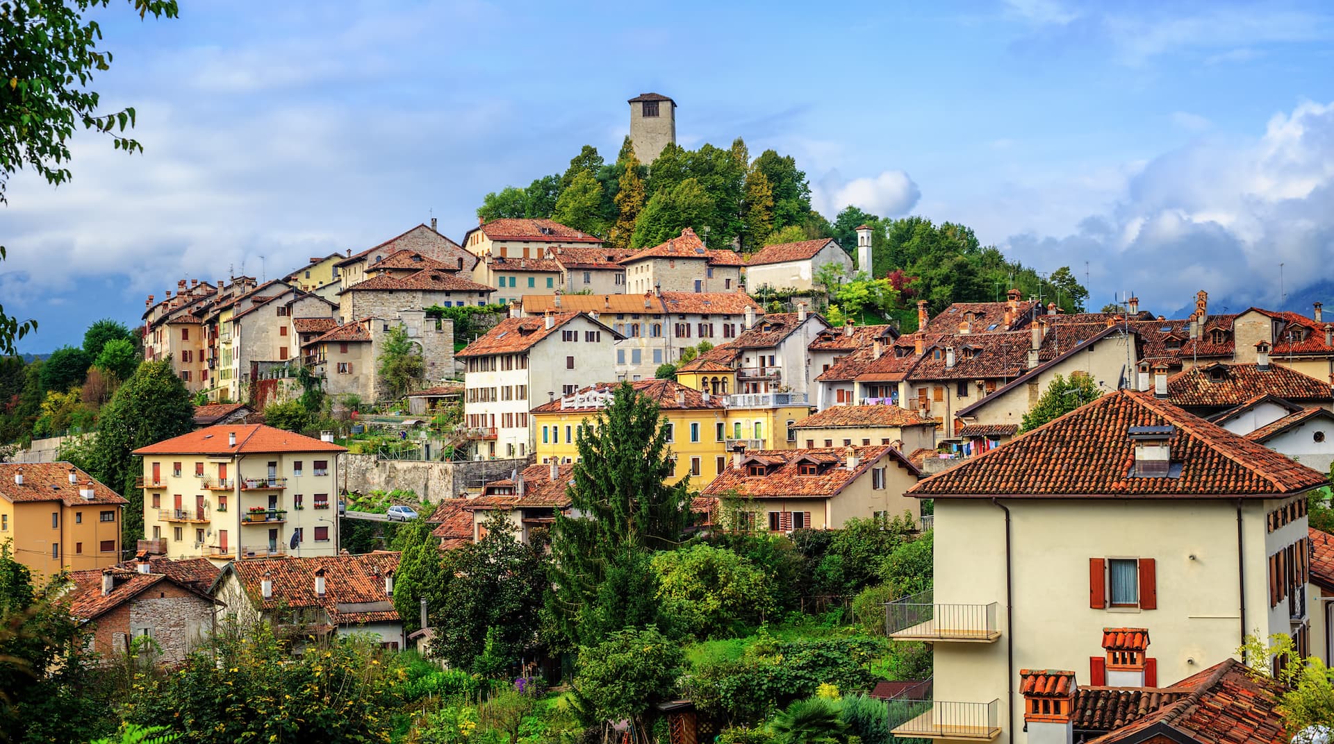 Hillside town with terracotta roofs, lush greenery, and a stone tower under a cloudy blue sky.