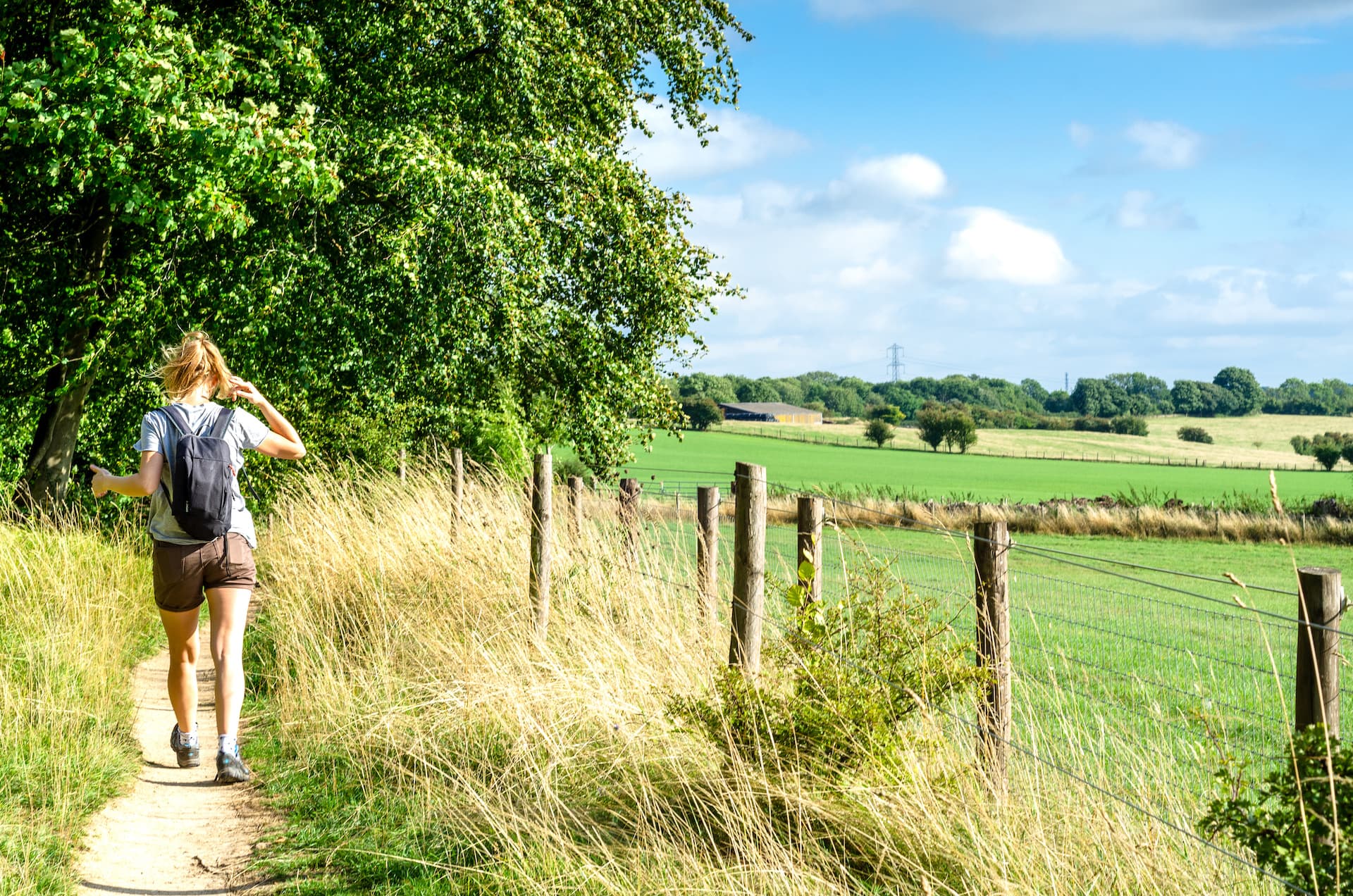 Woman hiking on dirt path beside tall grass and wooden fence toward green fields under blue sky.