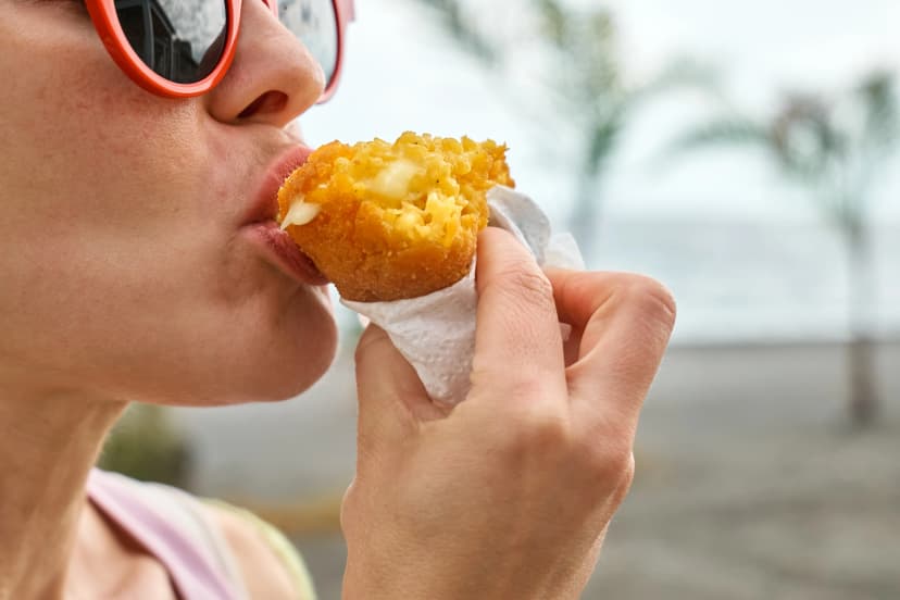 Young blond woman at seaside eating hot palatable arancini (deep fried rice balls with meat). Typical Sicilian street food. Close up.