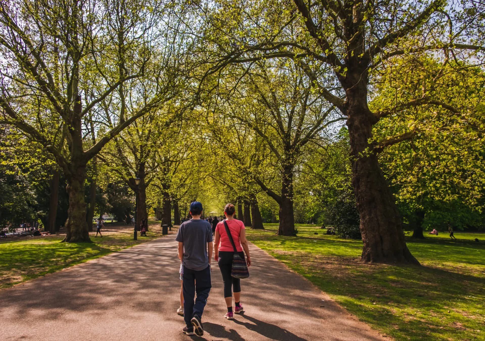 Woman and son walking down path under spring trees in city park with green grass.