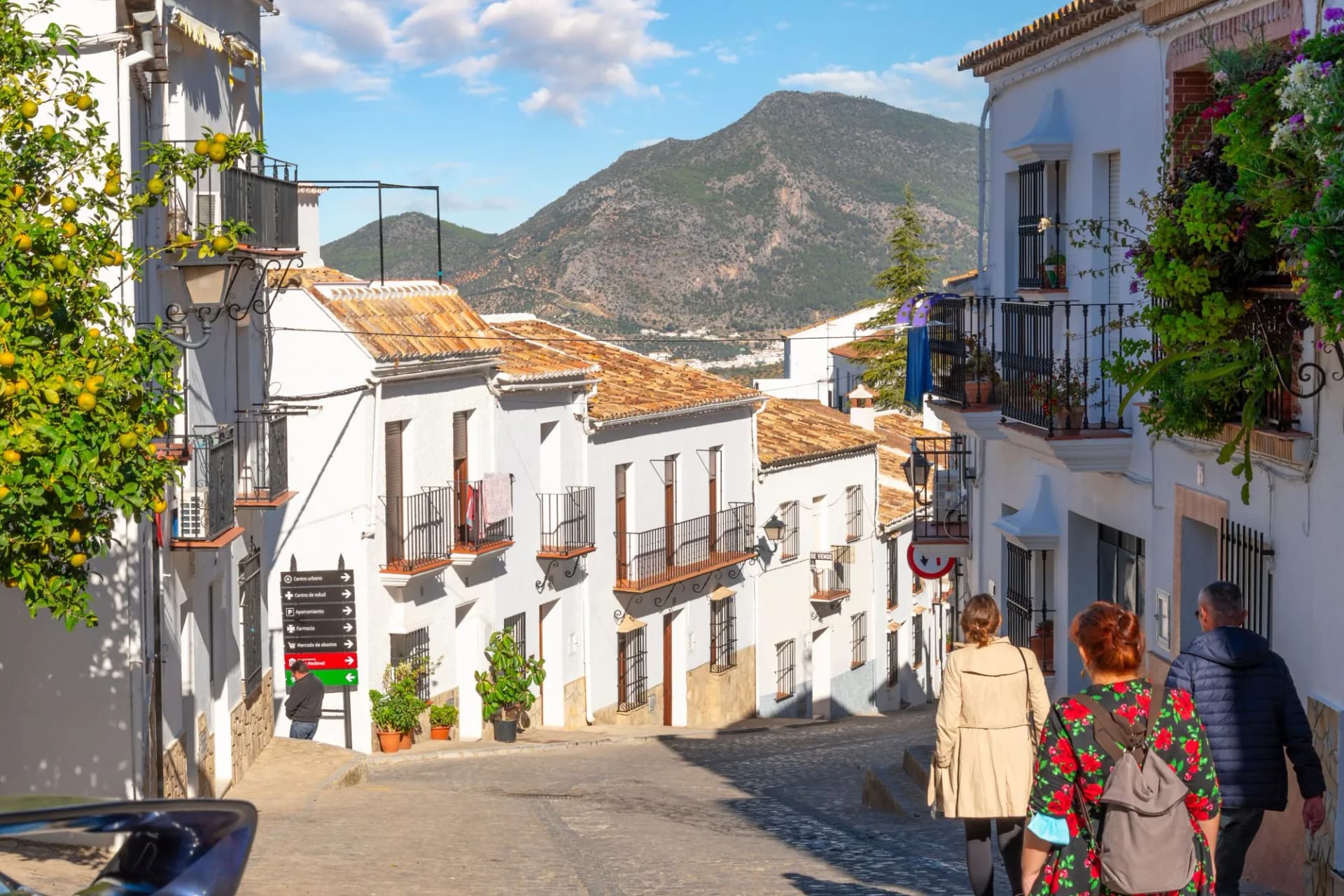 People walk up a cobblestone street lined with white Andalusian homes toward a large mountain in Zahara de la Sierra, Spain.