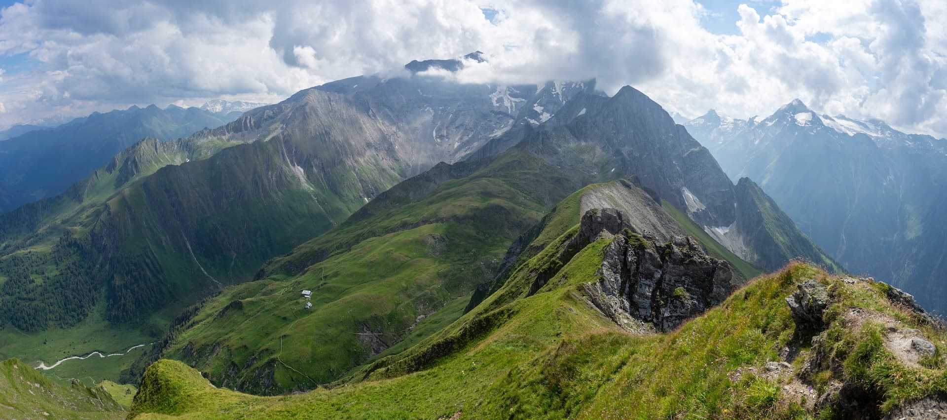 Alpine mountain ridge with green slopes, rocky outcrops, and snow-capped peaks under cloudy sky