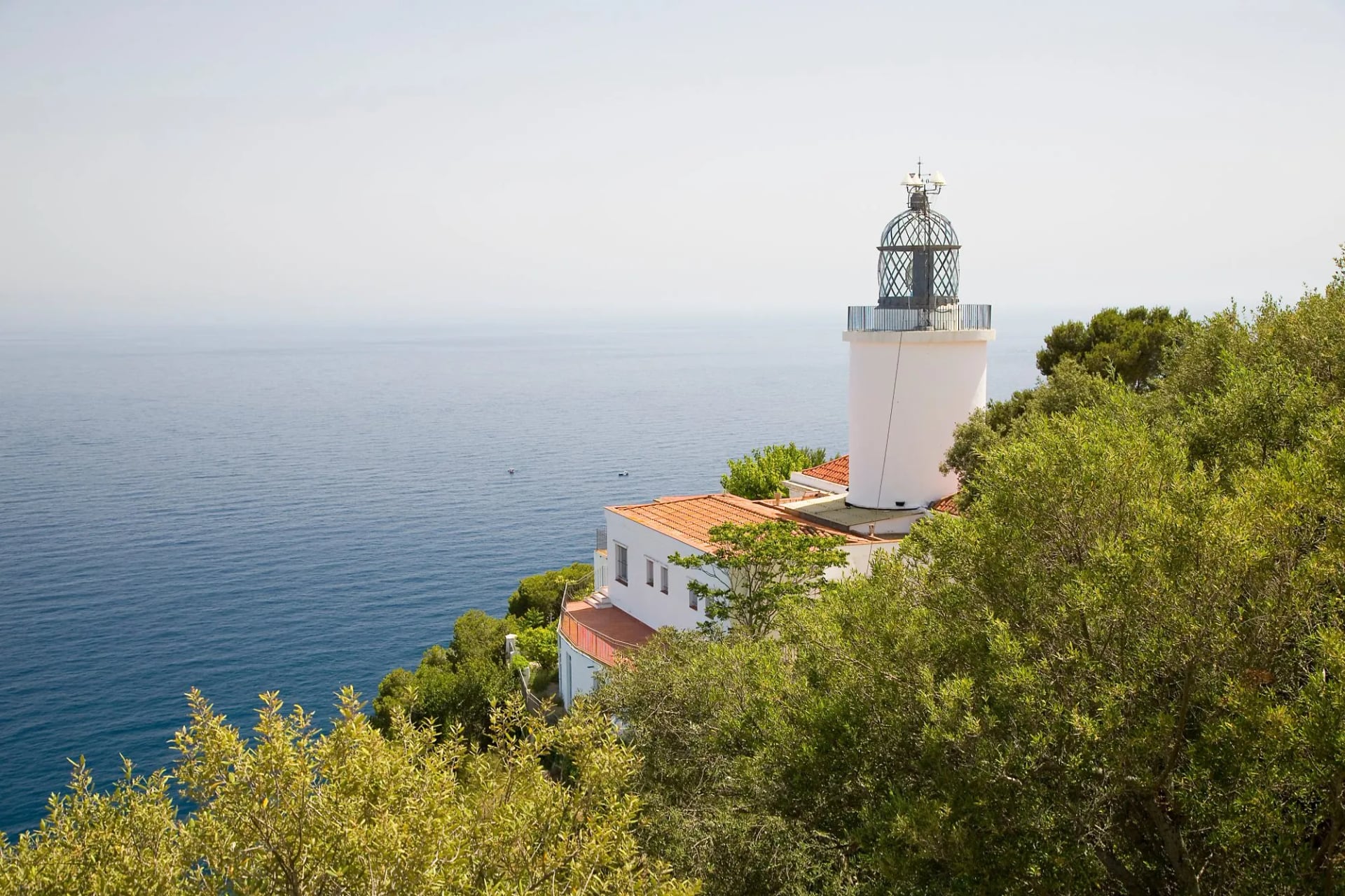 White lighthouse tower on the wooded Sant Sebastià headland above a calm sea