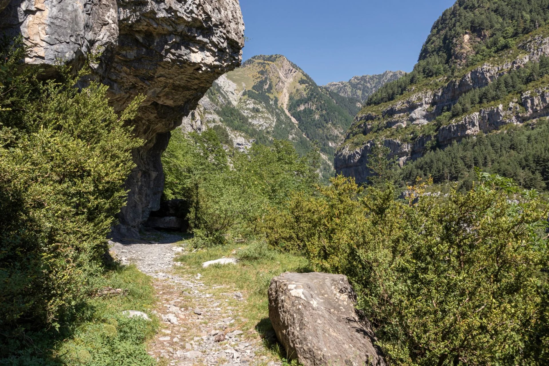 Walker on a stony path passing under a natural rock arch below limestone peaks