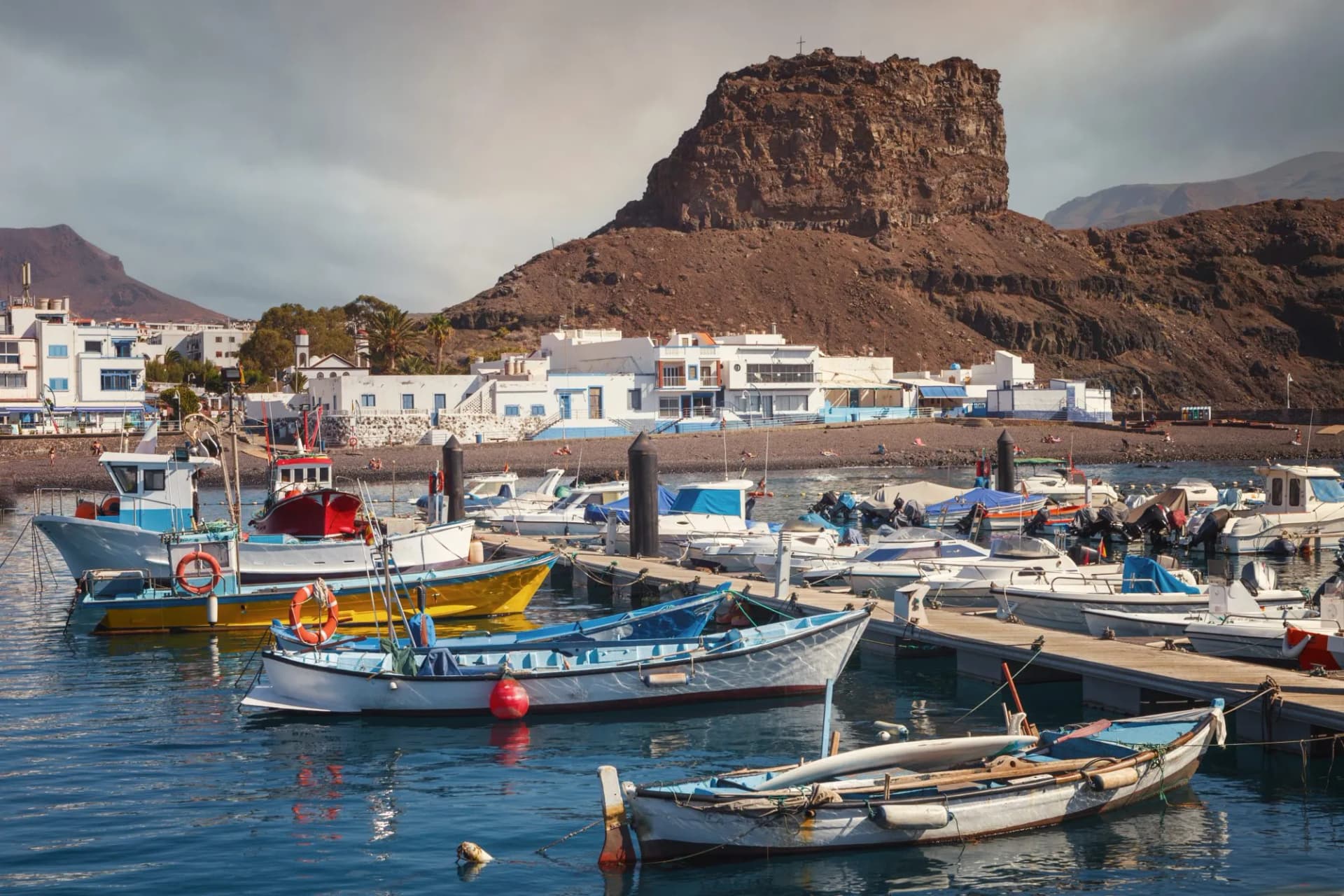 Boats moored in the port of Agaete, Gran Canaria, with white buildings and a large brown cliff backdrop.