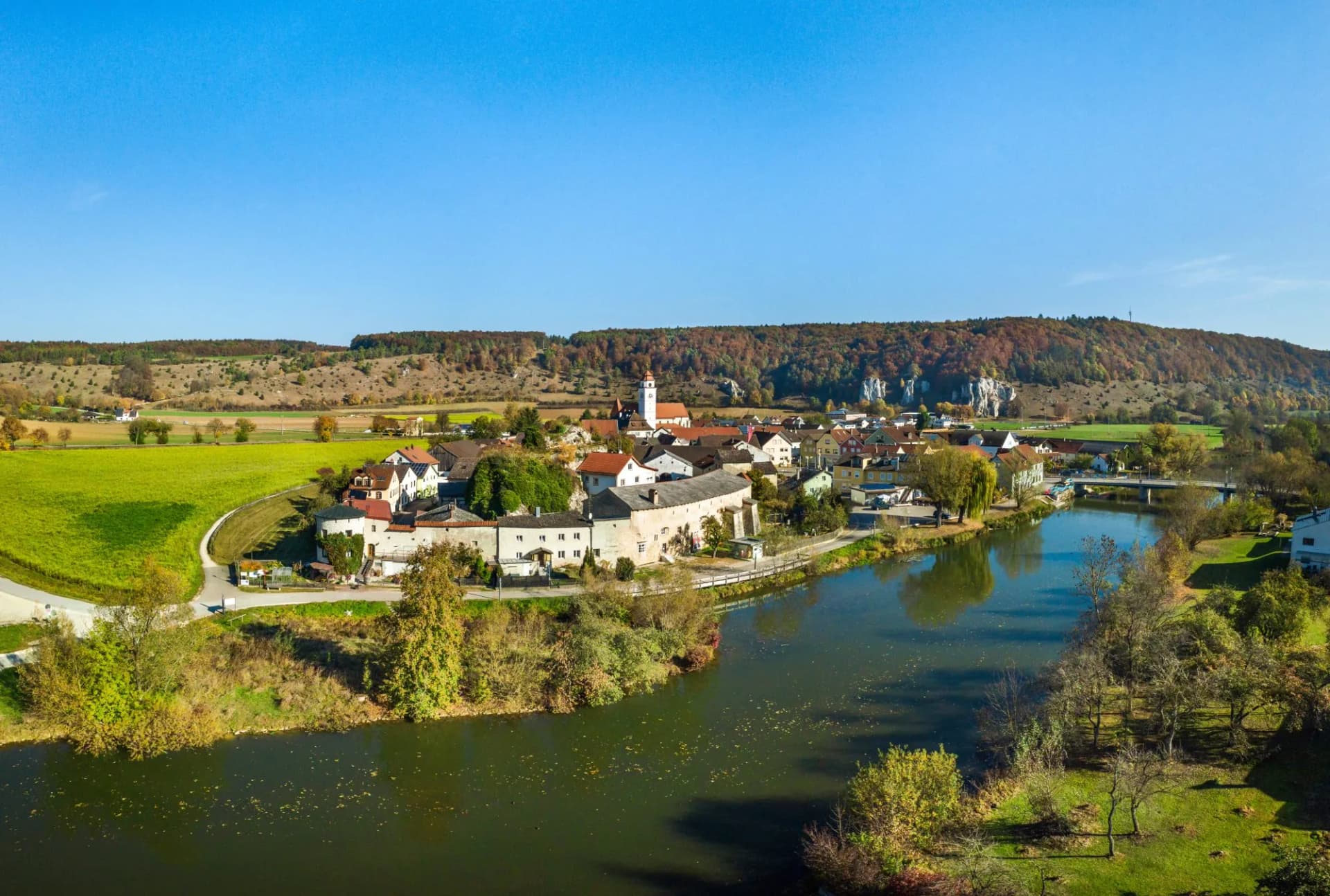 Village with church tower beside a river, set against rolling hills in autumn under a clear blue sky.