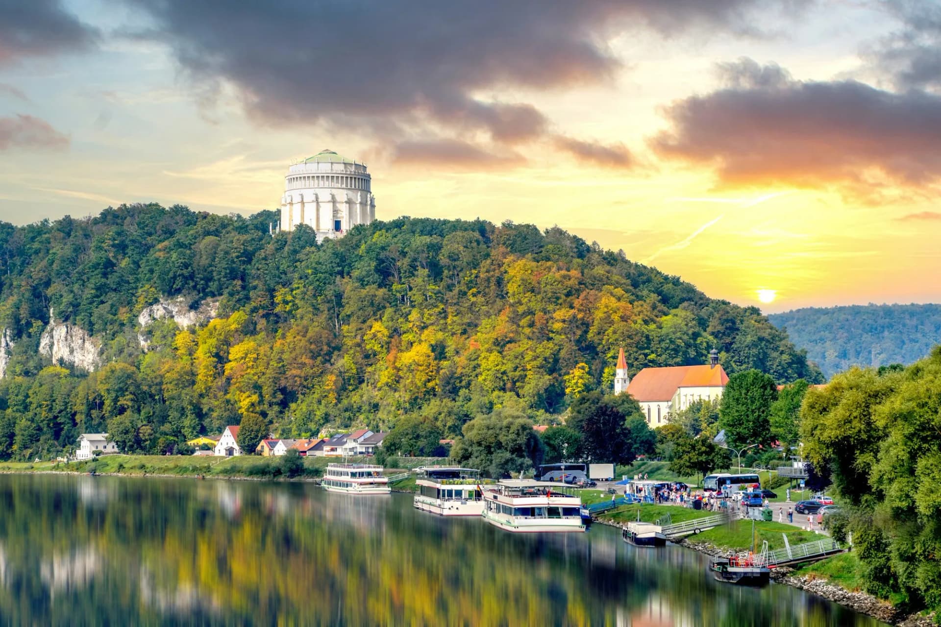 Walhalla monument above forested hill overlooking river with tour boats at sunset in Kelheim.