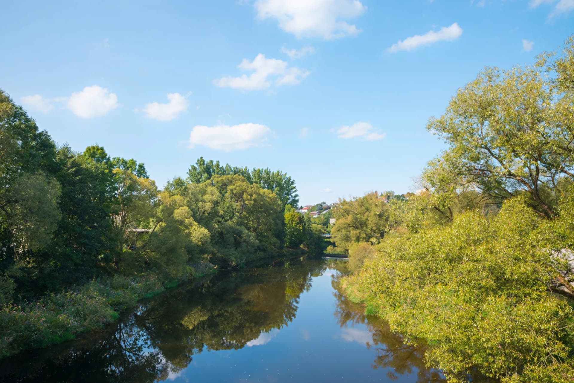 River flowing between lush green banks under a blue sky with scattered white clouds.