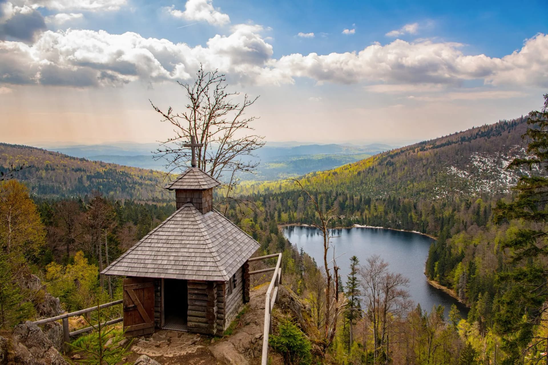 Wooden chapel overlooking Rachelsee lake surrounded by lush, rolling forested mountains under a cloudy sky.