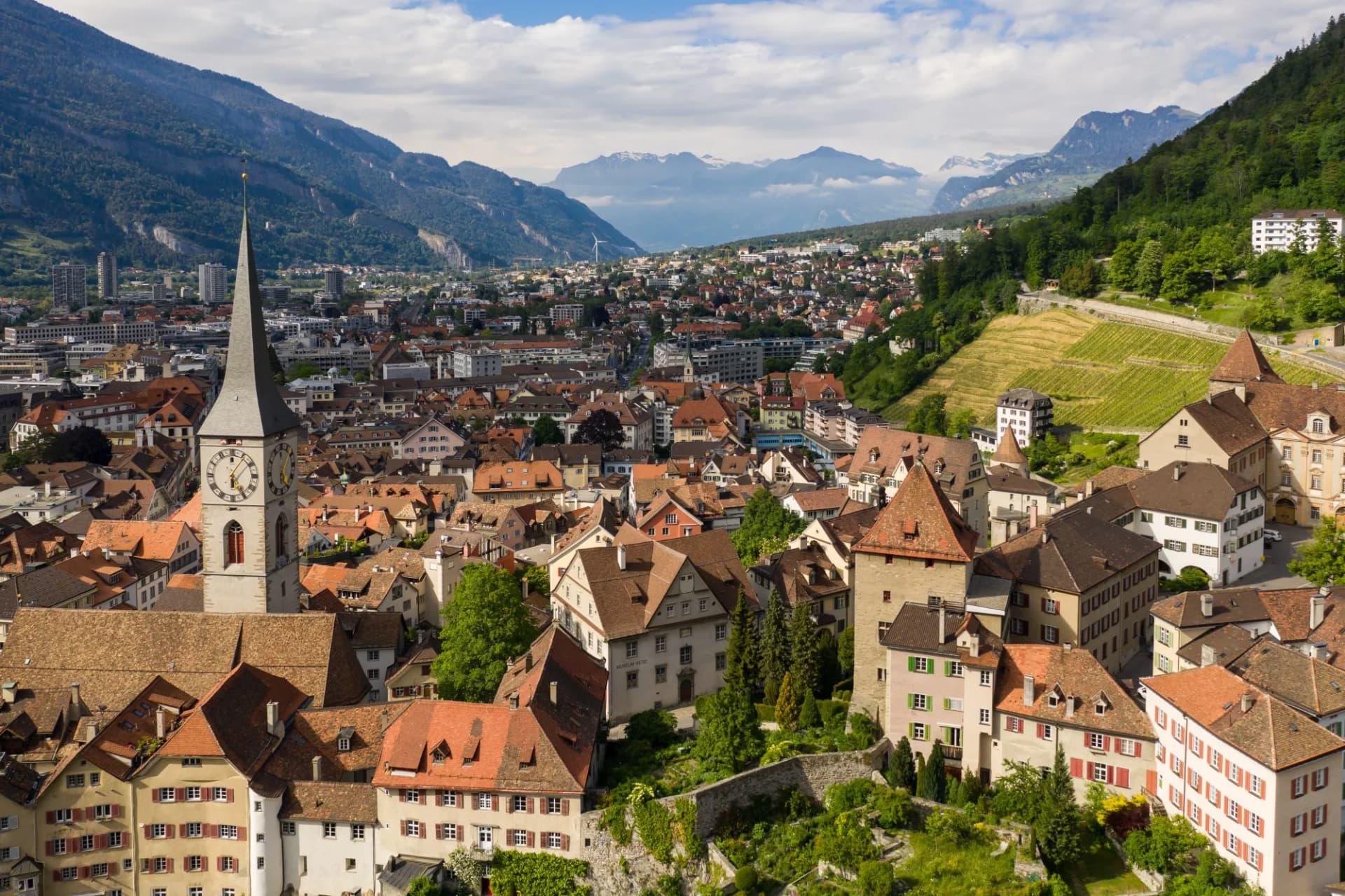 View of Chur city center with clock tower, terracotta roofs, and surrounding Alpine mountains.