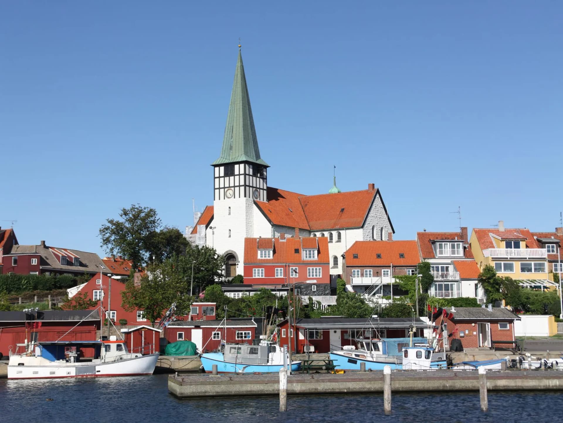 Fishing boats docked near colorful houses and a church with a tall green spire in Ronne.