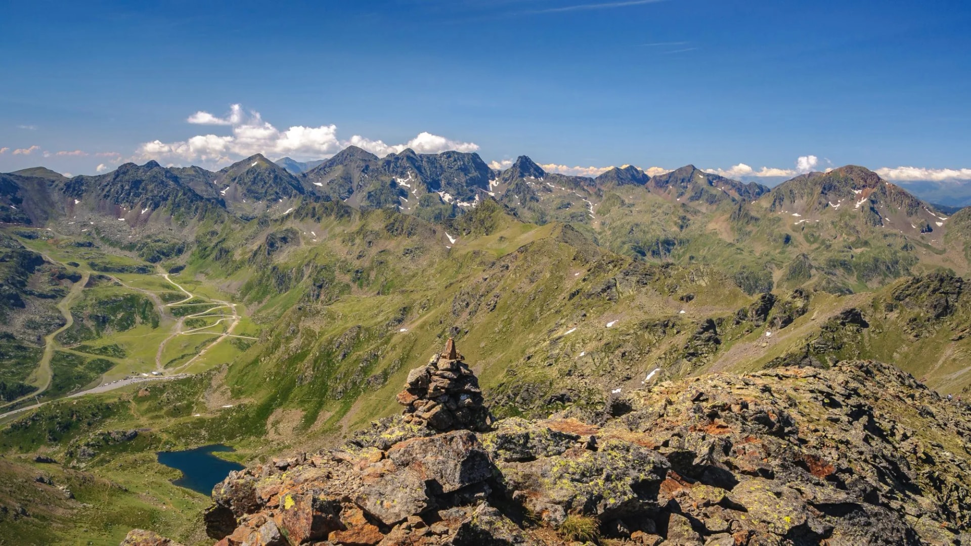 Rocky summit cairn overlooking green mountains, winding road, and small lake under blue sky.
