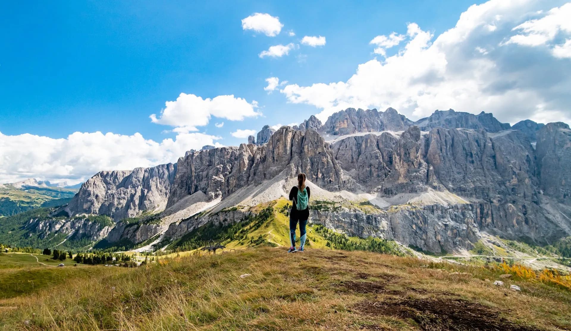 Hiker with backpack overlooking massive rocky mountains under blue sky near Gardena Pass.