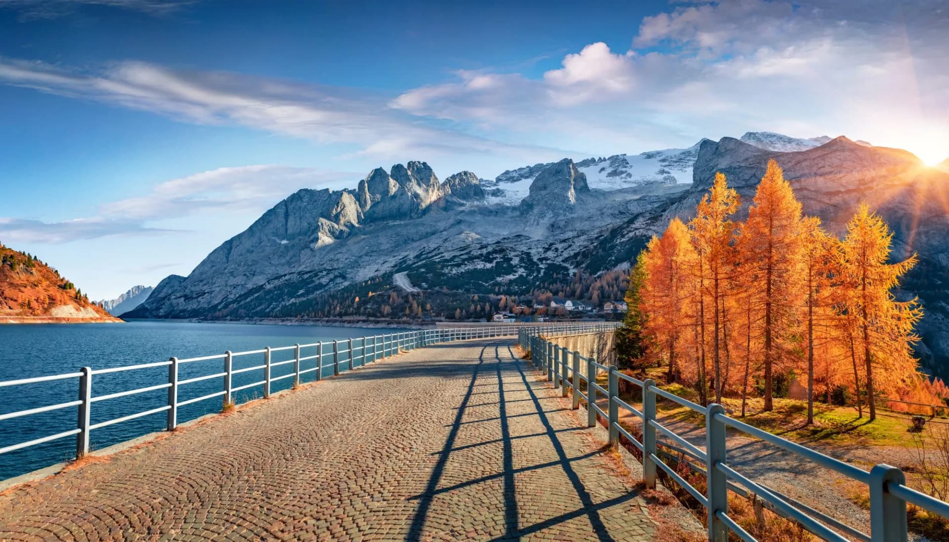Walkway on dam above Fedaia lake with majestic snowy mountains and autumn trees.