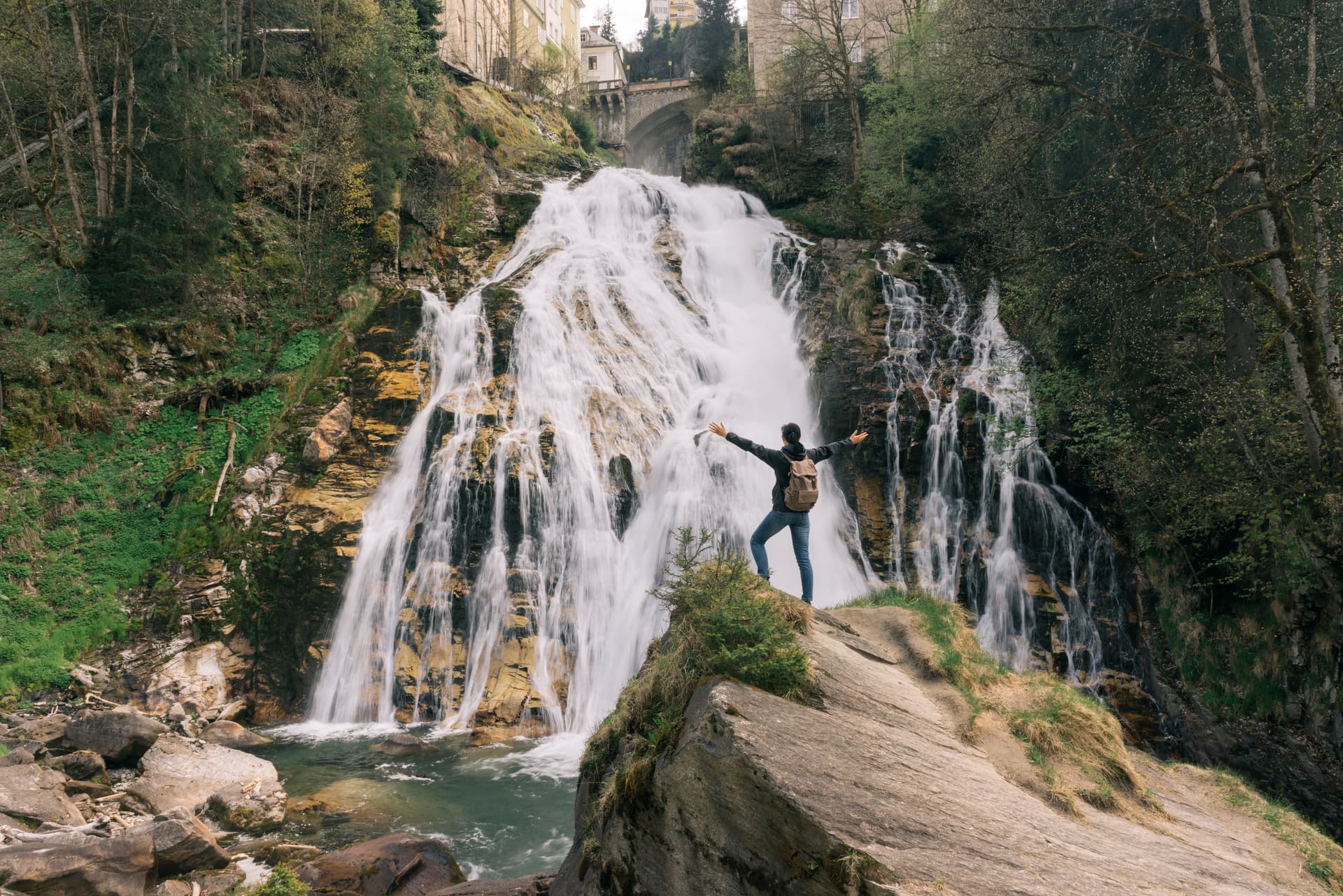 Hiker with arms spread facing large waterfall cascading down rocks near buildings in Austria.