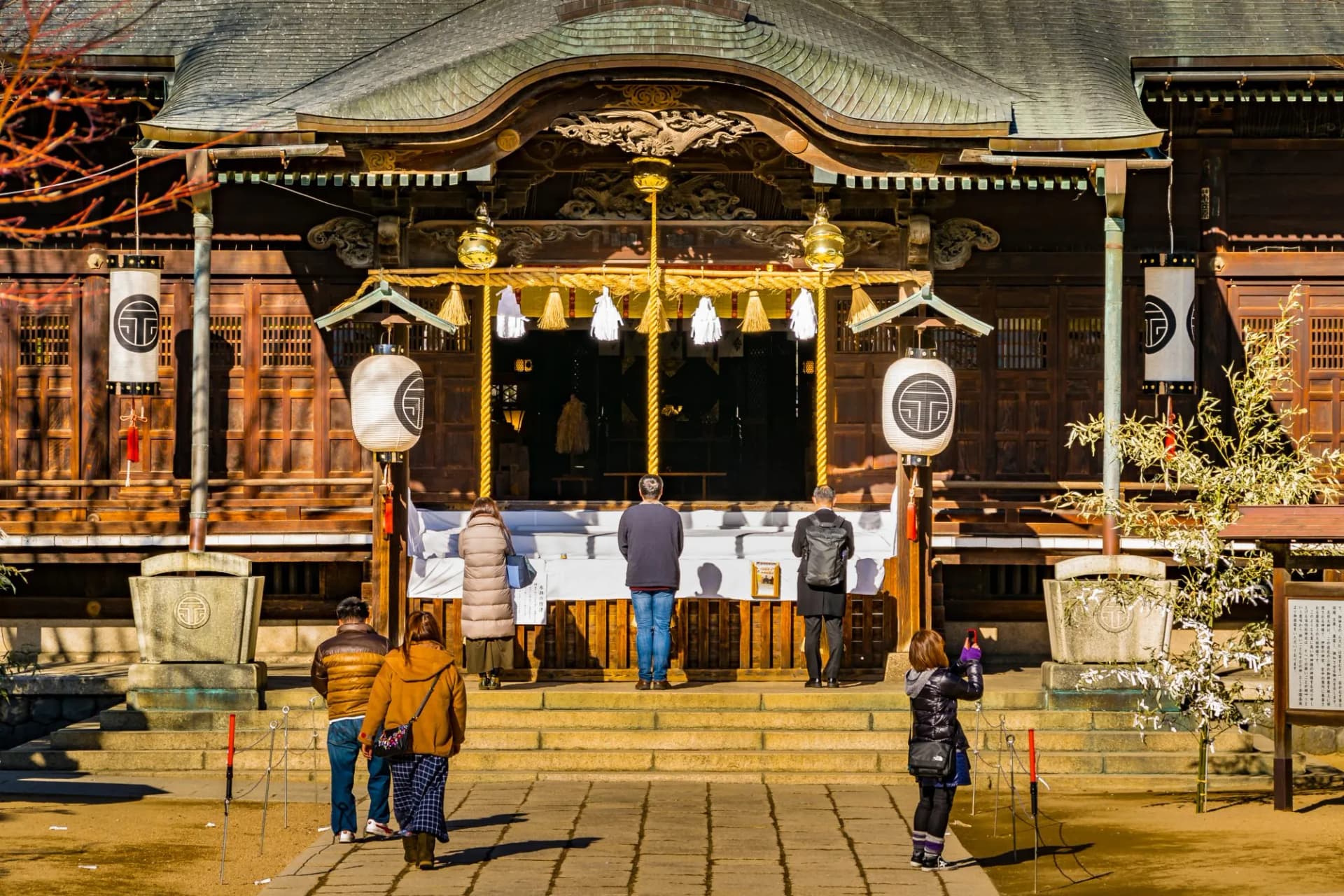 People praying at a traditional wooden Japanese shrine with lanterns and stone steps.