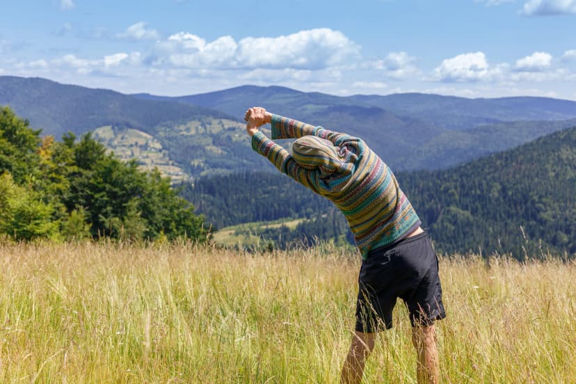 Back view of young man in green hoodie, exercising and stretching hands, looking at mountains range, resting after climbing and hiking on summer day - healthy lifestyle, travel, weekend trip concept