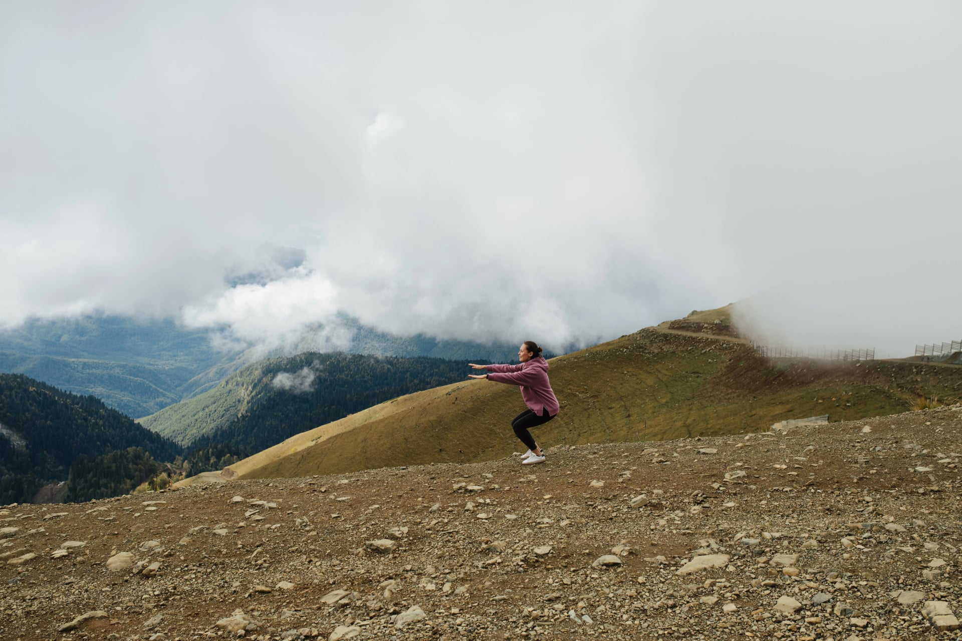 Physical exercises. Young woman doing squats high up in cloudy mountains.