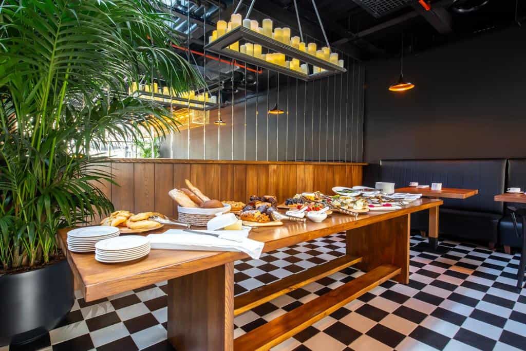 Breakfast buffet spread on a wooden table in a modern restaurant with checkered floors and indoor plants.