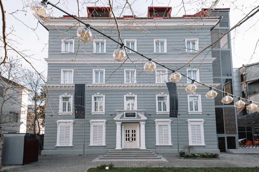 Gray multi-story building with white trim and string lights hanging in the foreground.