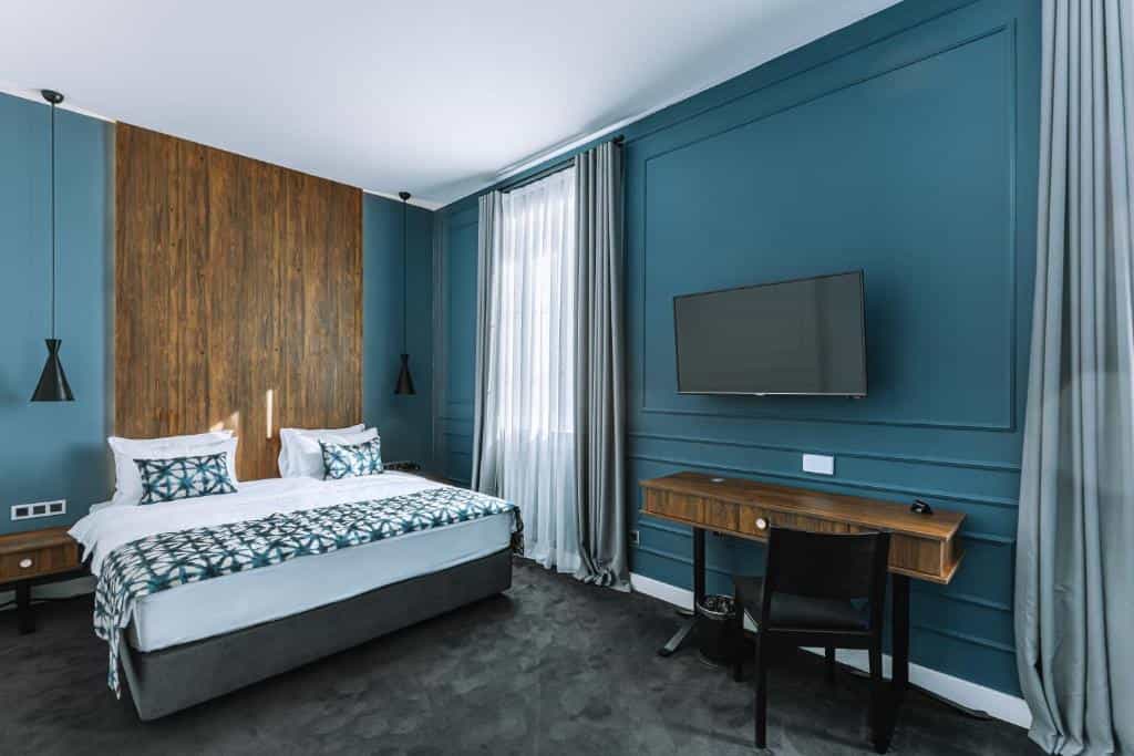 Hotel bedroom with a double bed, wooden accent wall, blue paneled walls, and a desk with a chair.